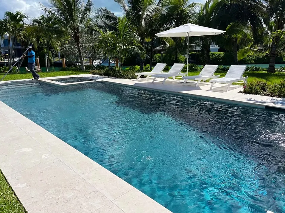 Waterfront swimming pool with sun loungers and a white umbrella on a stone deck beneath palm trees